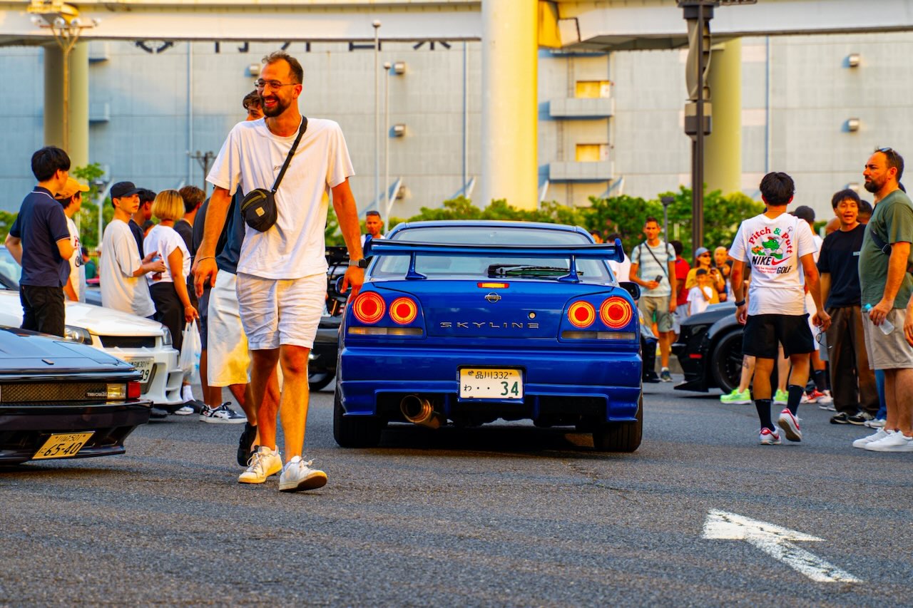 Rear of a blue Nissan Skyline GT-R R34 showing the SKYLINE badge at Daikoku PA at sunset