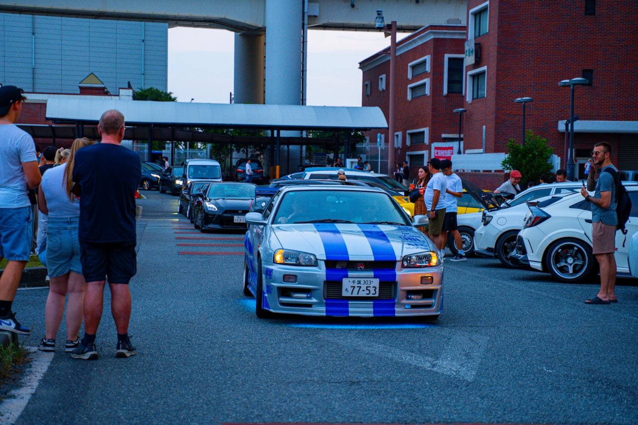 Striped Nissan Skyline R34 with blue underglow at Daikoku PA at dusk